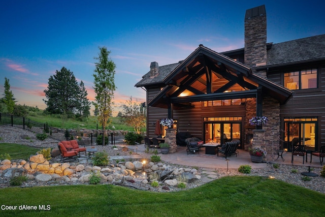 back of house at dusk with a patio, fence, a chimney, and stone siding