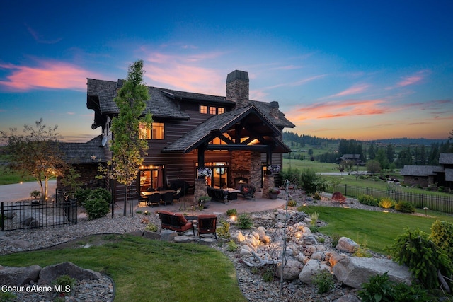 back of house at dusk featuring outdoor lounge area, fence, a chimney, a patio area, and a lawn