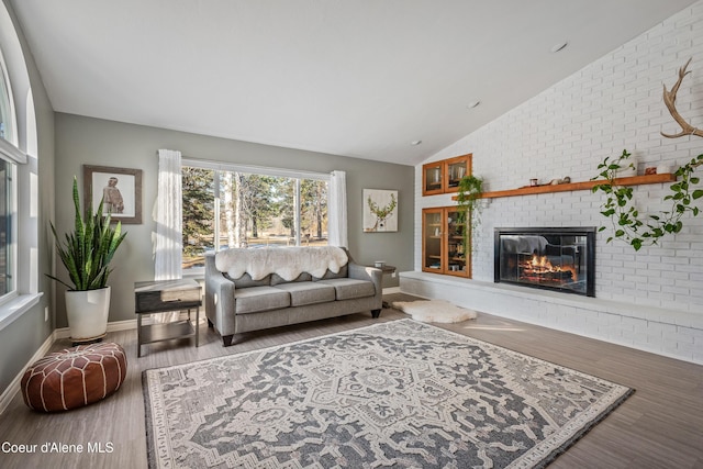 living room featuring wood finished floors, a fireplace, brick wall, baseboards, and lofted ceiling