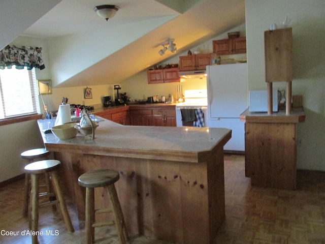 1959 Thimbleberry Ln, Sandpoint, ID 83864 - kitchen featuring brown cabinets, a kitchen breakfast bar, white appliances, under cabinet range hood, and vaulted ceiling