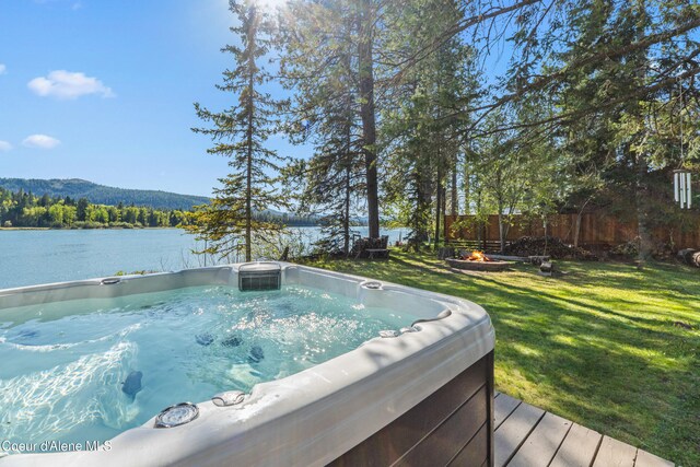 deck featuring a hot tub, a water view, an outdoor fire pit, and a view of trees