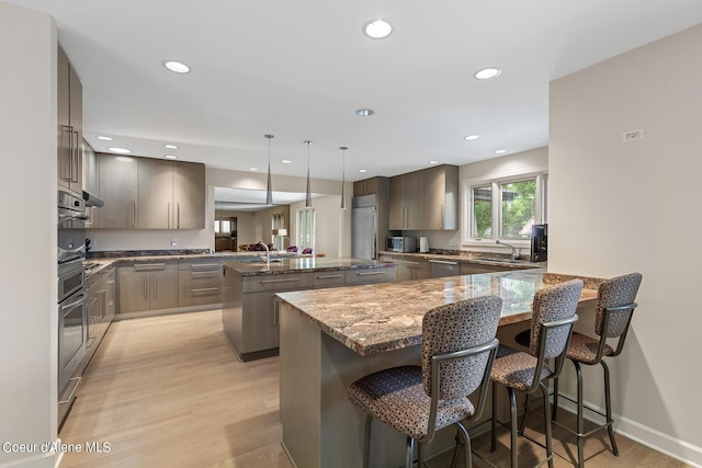 kitchen featuring an island with sink, light wood-style flooring, a peninsula, a sink, and stone counters