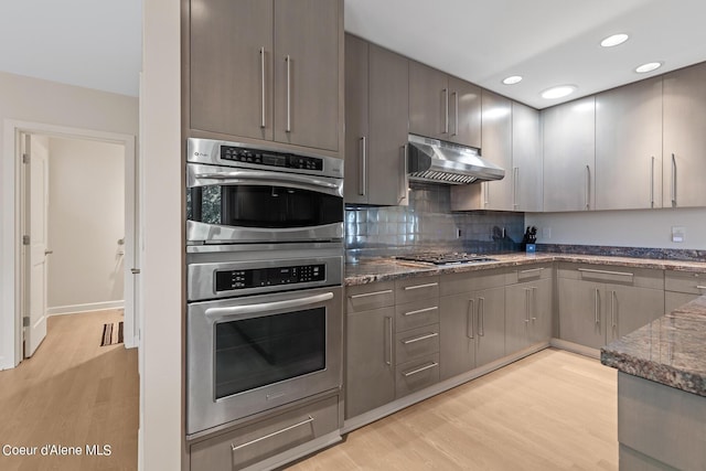 kitchen featuring under cabinet range hood, light wood finished floors, appliances with stainless steel finishes, dark stone counters, and backsplash