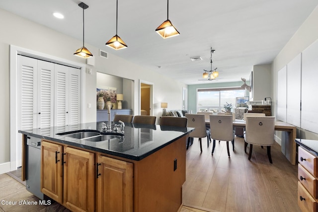 kitchen featuring a sink, dark stone counters, light wood-style flooring, a kitchen island with sink, and hanging light fixtures