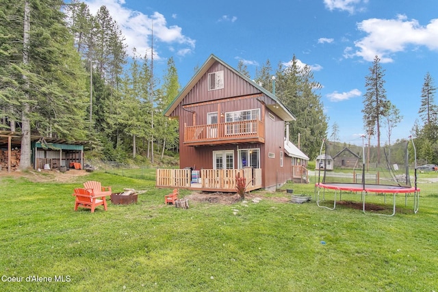 back of property featuring a trampoline, a wooden deck, a yard, a fire pit, and board and batten siding