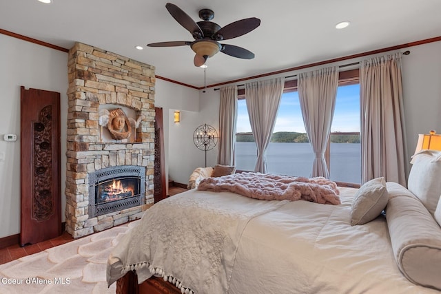 bedroom with crown molding, wood finished floors, a stone fireplace, a water view, and baseboards