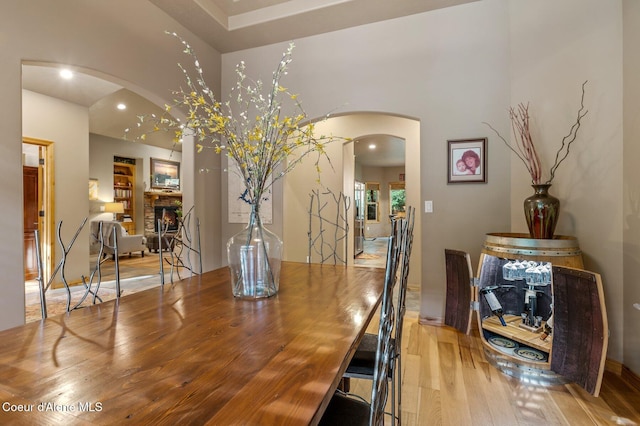 dining area with arched walkways, light wood-style floors, a stone fireplace, a towering ceiling, and recessed lighting