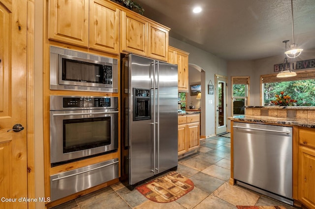 kitchen featuring stainless steel appliances, a warming drawer, arched walkways, decorative light fixtures, and dark stone countertops