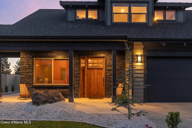 11387 N Armonia Way, Hayden Lake, ID 83835 - exterior entry at dusk featuring roof with shingles, stone siding, concrete driveway, an attached garage, and a porch