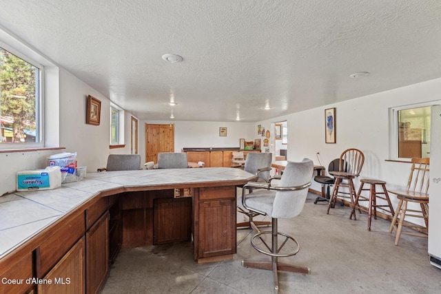 26277 N Chevrolane Way, Rathdrum, ID 83858 - kitchen featuring tile countertops, a textured ceiling, a peninsula, brown cabinetry, and concrete flooring