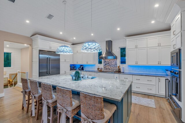 kitchen with wall chimney range hood, decorative light fixtures, built in appliances, and a spacious island