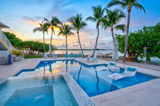 pool at dusk featuring a patio area, an in ground hot tub, and a water view