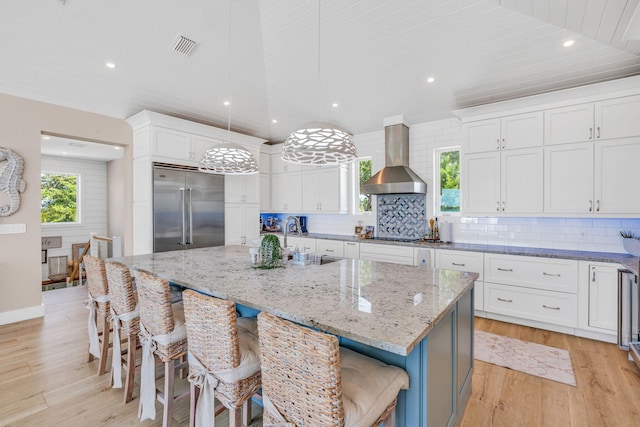 kitchen with white cabinetry, wall chimney exhaust hood, stainless steel built in fridge, decorative light fixtures, and a large island with sink