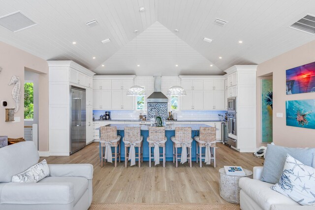 kitchen featuring a kitchen island with sink, built in appliances, a kitchen bar, decorative backsplash, and wall chimney exhaust hood