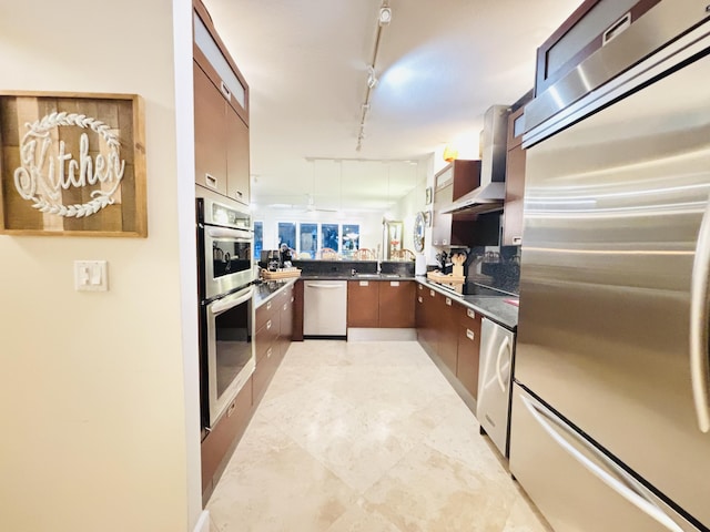 kitchen featuring dark countertops, track lighting, stainless steel appliances, wall chimney exhaust hood, and a sink