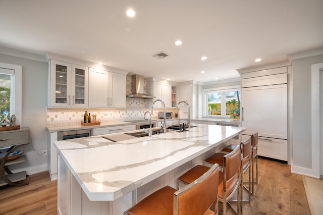 kitchen with wall chimney exhaust hood, light stone counters, a kitchen breakfast bar, an island with sink, and white cabinets