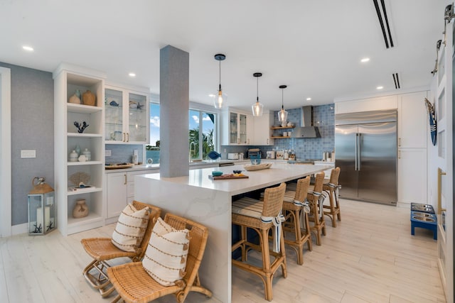 kitchen featuring white cabinetry, a center island, built in fridge, pendant lighting, and wall chimney range hood