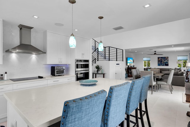 kitchen featuring wall chimney exhaust hood, black electric cooktop, double oven, pendant lighting, and white cabinets