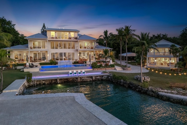 back house at dusk with pool water feature, a water view, a balcony, a yard, and a patio