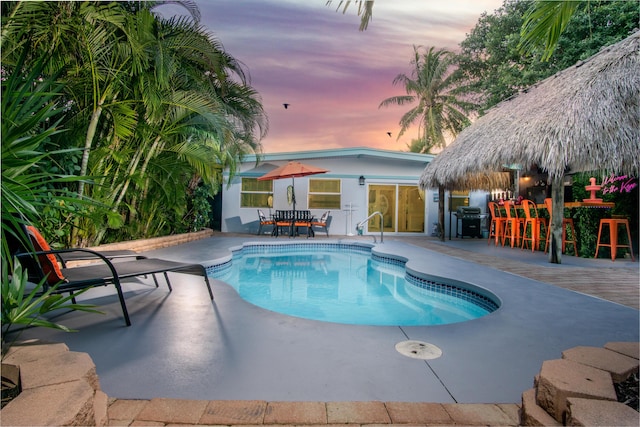 pool at dusk featuring an outdoor bar, grilling area, and a patio area