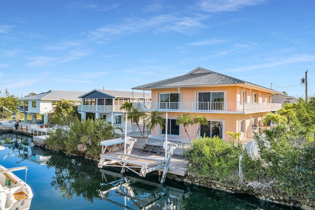 rear view of house with stucco siding, a water view, a balcony, metal roof, and boat lift