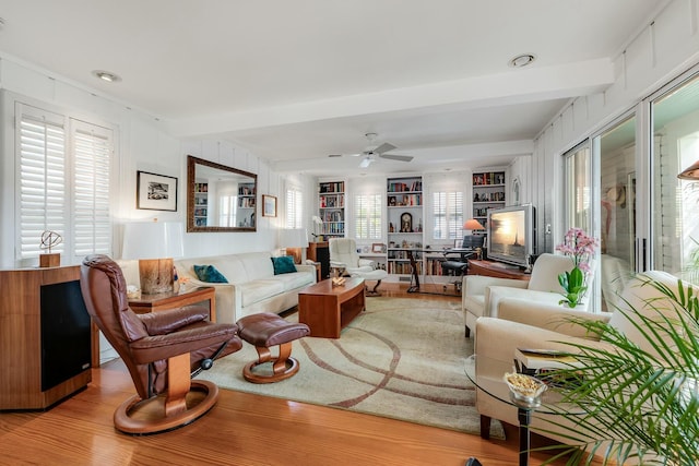 living room with built in shelves, beam ceiling, ceiling fan, and light wood finished floors