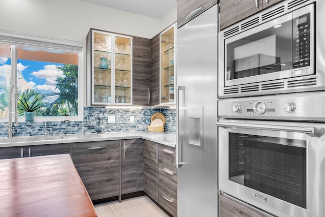 kitchen with tasteful backsplash, sink, built in appliances, light tile patterned floors, and dark brown cabinets