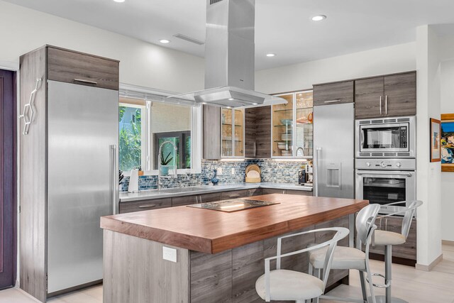 kitchen featuring dark brown cabinets, a center island, built in appliances, tasteful backsplash, and island exhaust hood