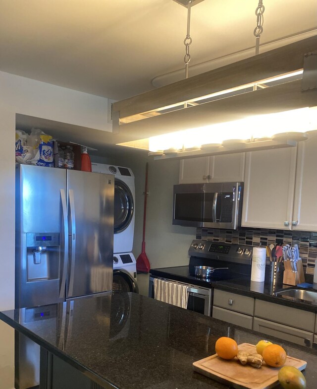 kitchen featuring stacked washer / drying machine, white cabinetry, dark stone countertops, appliances with stainless steel finishes, and decorative backsplash