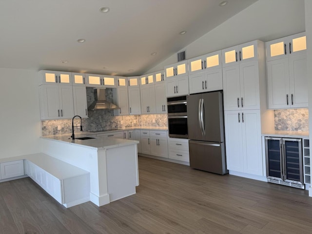 kitchen featuring a peninsula, a sink, vaulted ceiling, appliances with stainless steel finishes, and wall chimney exhaust hood