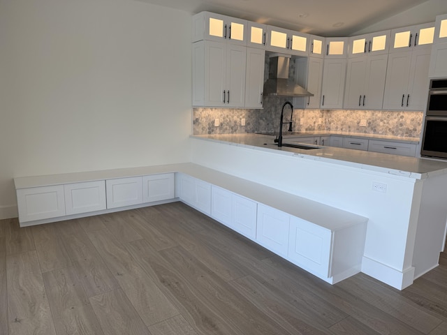 kitchen featuring backsplash, a peninsula, wood finished floors, stainless steel double oven, and wall chimney exhaust hood