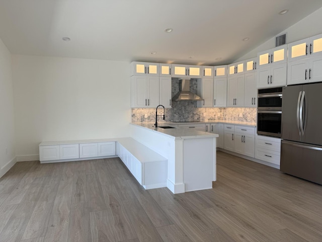 kitchen featuring a peninsula, a sink, appliances with stainless steel finishes, wall chimney range hood, and tasteful backsplash