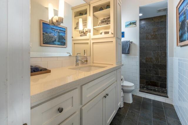 full bathroom with toilet, tile walls, tiled shower, vanity, and wainscoting
