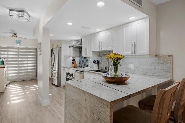 kitchen with wall chimney range hood, sink, stainless steel appliances, white cabinets, and kitchen peninsula