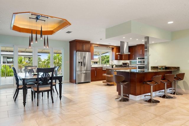 kitchen featuring a breakfast bar, island range hood, appliances with stainless steel finishes, a tray ceiling, and kitchen peninsula