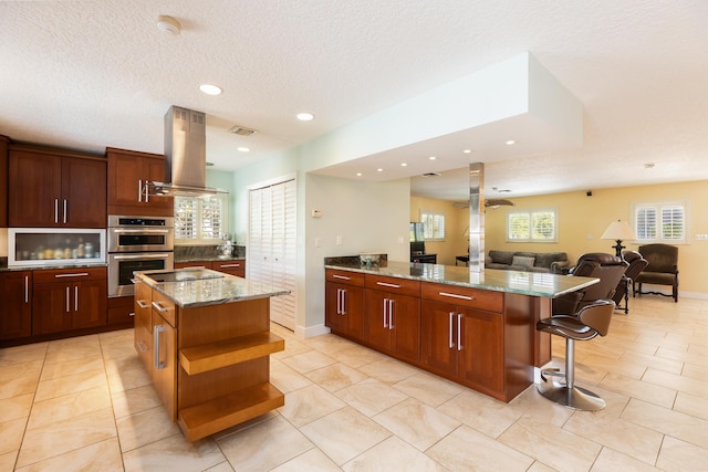 kitchen with island exhaust hood, a center island, double oven, and light stone countertops