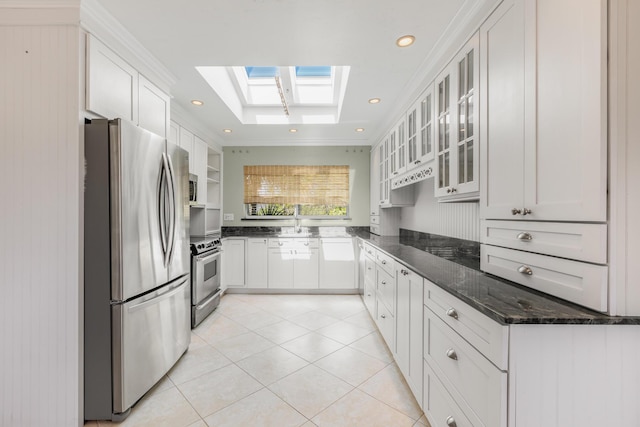 kitchen featuring light tile patterned floors, appliances with stainless steel finishes, white cabinetry, dark stone countertops, and a skylight