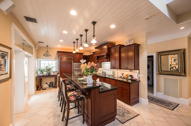 kitchen featuring decorative light fixtures, a breakfast bar area, a kitchen island with sink, stacked washer / drying machine, and wood ceiling