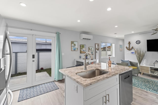 kitchen with a wall unit AC, a kitchen island with sink, a sink, white cabinetry, and appliances with stainless steel finishes