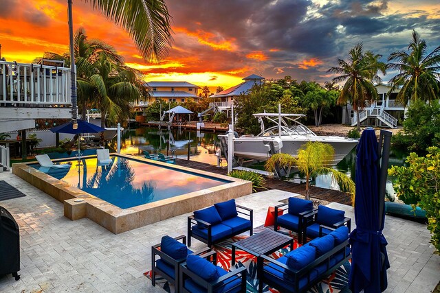 pool at dusk with a water view, outdoor lounge area, a patio area, and a boat dock