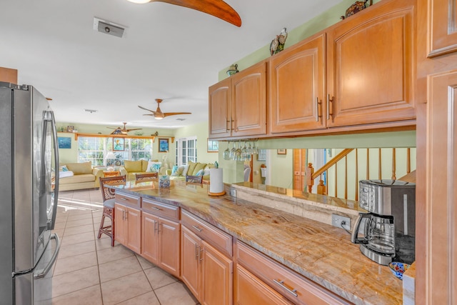 kitchen with light tile patterned flooring, kitchen peninsula, stainless steel refrigerator, light stone counters, and ceiling fan