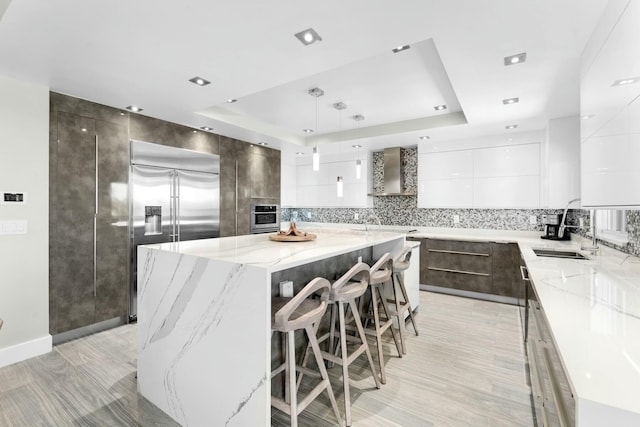 kitchen with a spacious island, a raised ceiling, stainless steel appliances, wall chimney exhaust hood, and modern cabinets