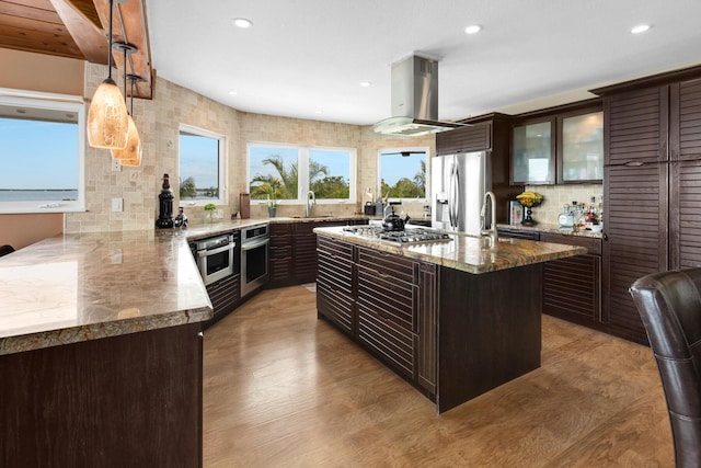 kitchen with island range hood, hanging light fixtures, dark stone countertops, appliances with stainless steel finishes, and an island with sink