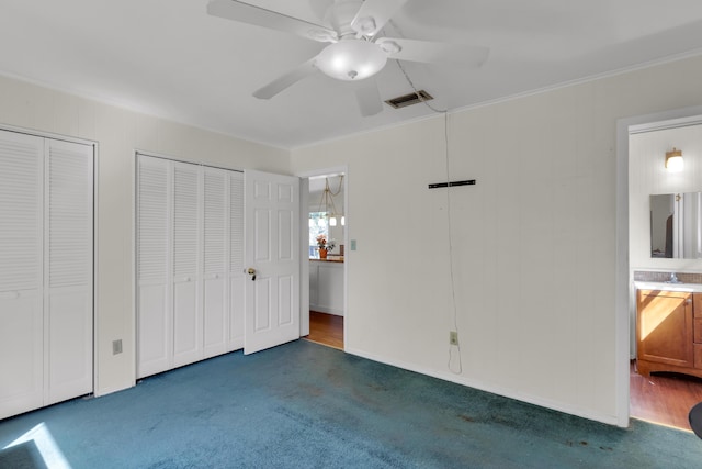 unfurnished bedroom with dark colored carpet, ensuite bath, ornamental molding, two closets, and ceiling fan