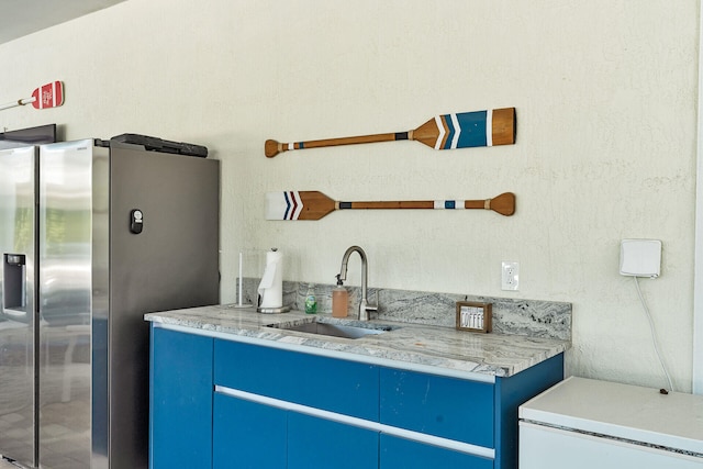 kitchen with stainless steel fridge with ice dispenser, blue cabinetry, sink, and light stone countertops