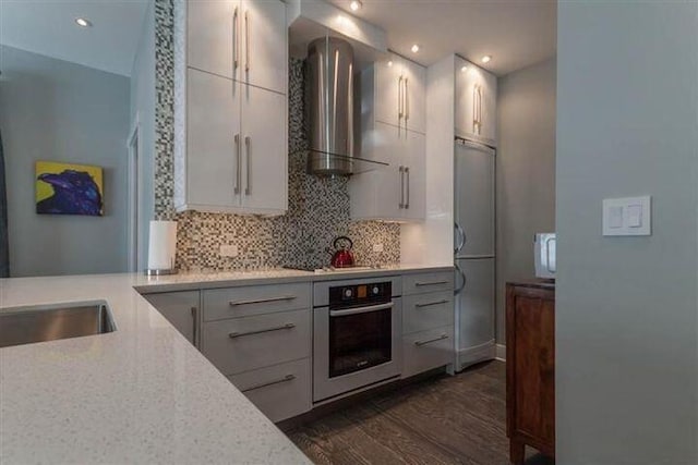 kitchen featuring wall chimney range hood, dark wood-type flooring, white cabinets, decorative backsplash, and oven