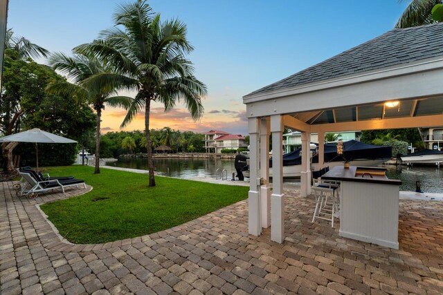 patio terrace at dusk featuring a water view, a gazebo, an outdoor bar, and a lawn