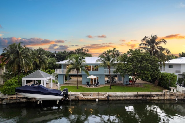 back house at dusk with a water view, a balcony, and a yard