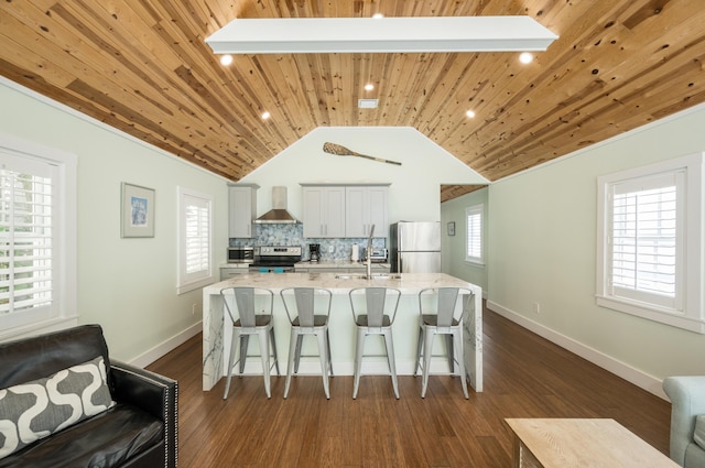 kitchen featuring wall chimney range hood, appliances with stainless steel finishes, a kitchen island with sink, a kitchen breakfast bar, and light stone counters