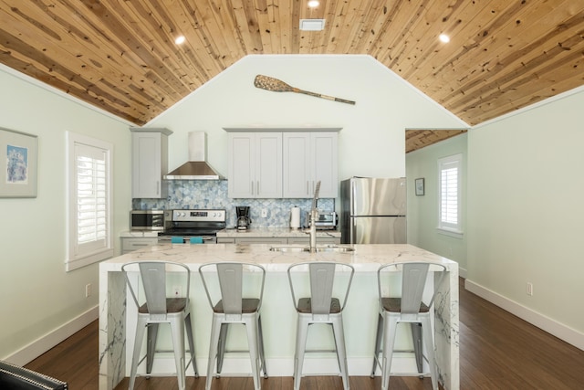 kitchen featuring white cabinetry, appliances with stainless steel finishes, an island with sink, light stone countertops, and wall chimney range hood